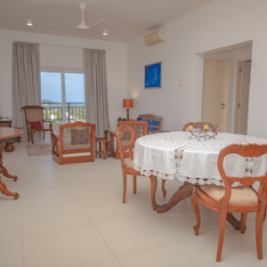 A spacious living area in Aganthuka Villas, with comfortable seating, a large dining table, and a flat-screen TV mounted on a Dutch box, overlooking the ocean through floor-to-ceiling windows. The room is decorated in neutral tones with blue accents and contemporary furnishings.