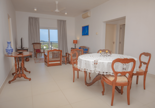 A spacious living area in Aganthuka Villas, with comfortable seating, a large dining table, and a flat-screen TV mounted on a Dutch box, overlooking the ocean through floor-to-ceiling windows. The room is decorated in neutral tones with blue accents and contemporary furnishings.
