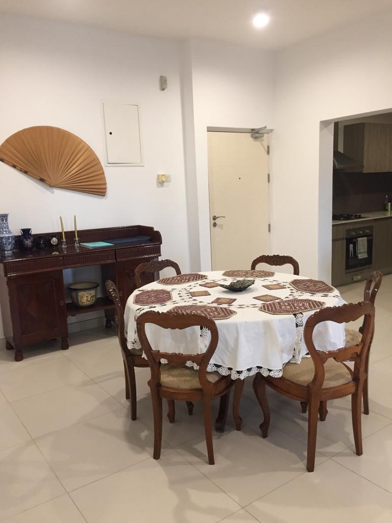 View from the dining area of Aganthuka Galle towards the main entrance, featuring a large round table set with six chairs, white tablecloth, and table mats. An antique buffet table stands against the wall, adding a touch of elegance. A glimpse into the kitchen reveals the continuity of the open and inviting space.