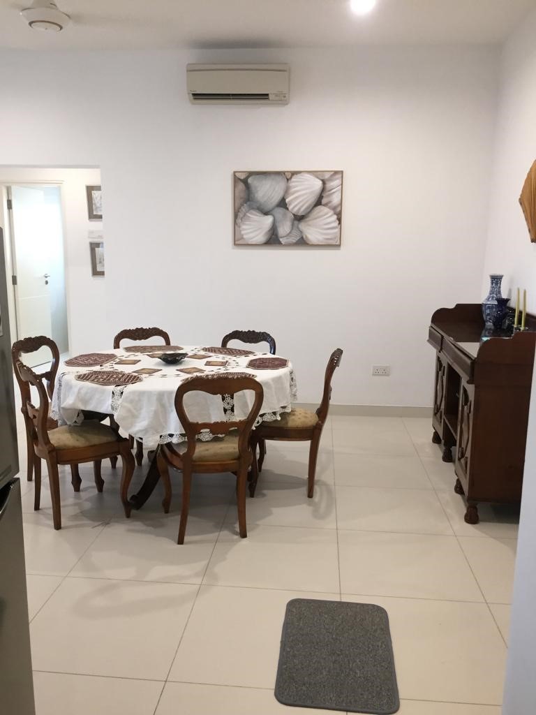 View from the kitchen at Aganthuka Galle, looking towards the common toilet. The scene includes a large round table set with six chairs, covered with a white tablecloth and table mats, creating a welcoming dining space. An antique buffet table against the wall enhances the room's elegance. This perspective highlights the cohesive and sophisticated interior design of the living space.