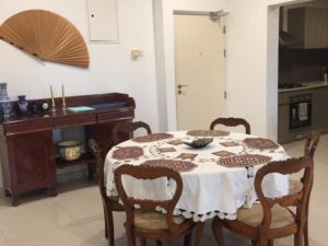 View from the dining area of Aganthuka Galle towards the main entrance, featuring a large round table set with six chairs, white tablecloth, and table mats. An antique buffet table stands against the wall, adding a touch of elegance. A glimpse into the kitchen reveals the continuity of the open and inviting space.