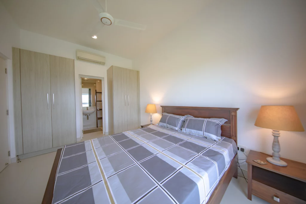 Elegant master bedroom at Aganthuka Galle featuring wall-to-ceiling cupboards, with a view into the ensuite. The room is equipped with air conditioning and a ceiling fan, ensuring comfort. Two bedside tables with lamps provide ambient lighting, creating a warm and inviting atmosphere.